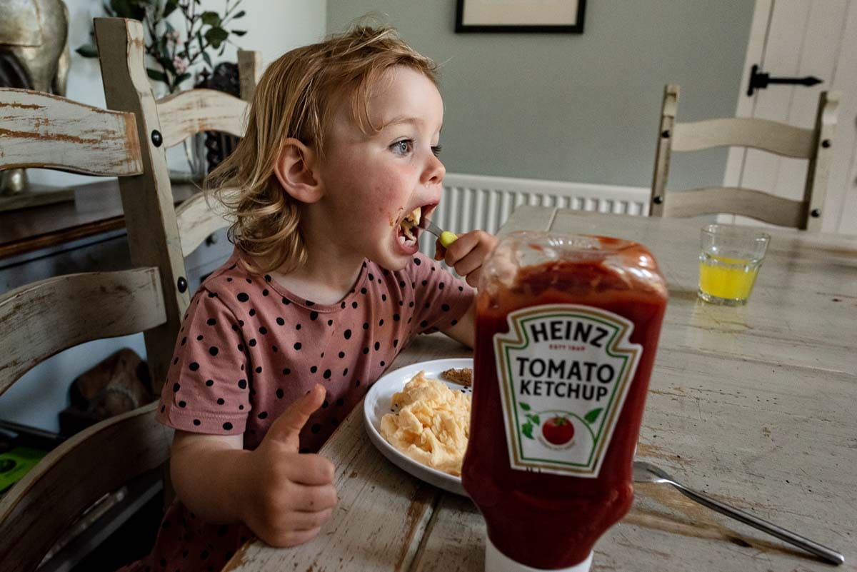 child at the dinner table eating scrambled egg with large bottle of ketchup